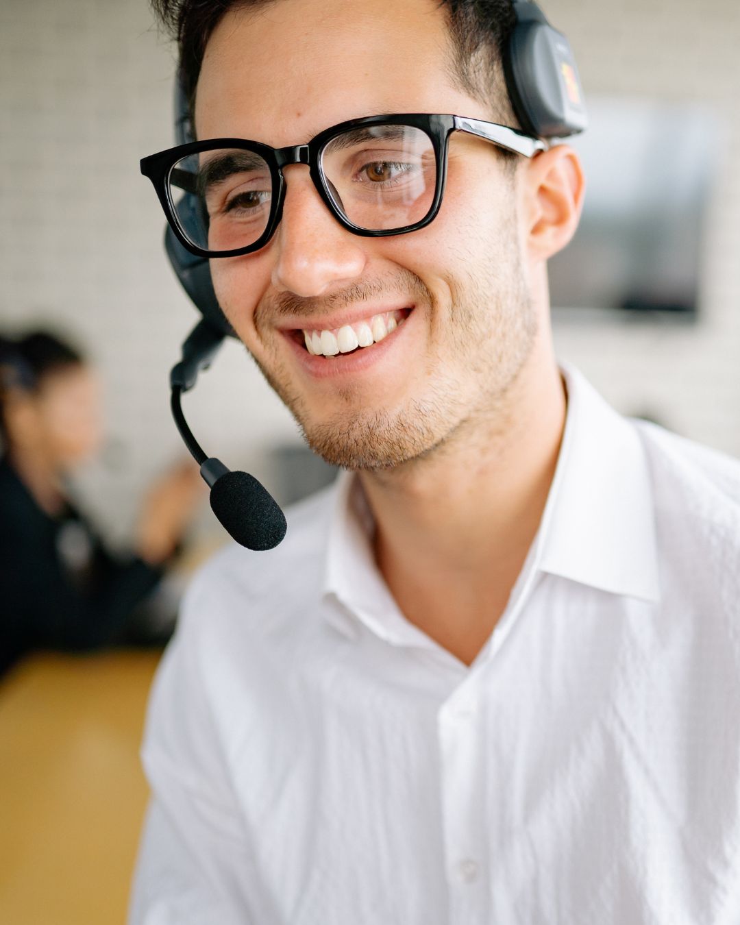 A man wearing glasses and a headset smiles while sitting indoors, dressed in a white shirt.